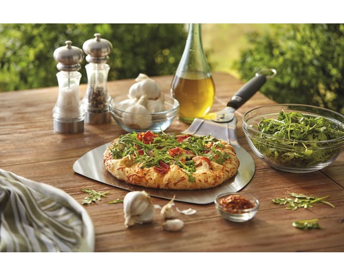 Scenă cu pizza pe o lopată de pizza, usturoi, rucola, ulei, condimente și tăietor de pizza pe o masă de lemn.