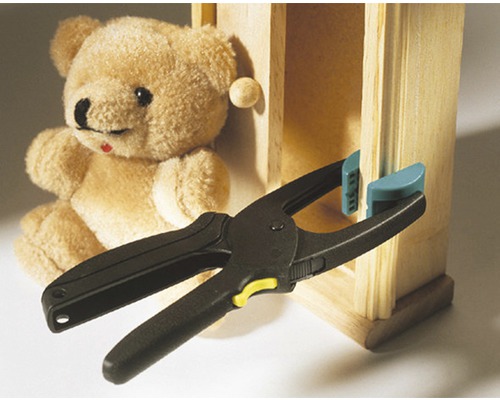 Clește cu arc care fixează o bucată de lemn de un dulap miniatural, cu un ursuleț de pluș în fundal
