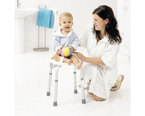 O femeie stă în genunchi lângă un copil așezat pe un scaun de duș cu brațe într-o baie luminoasă.