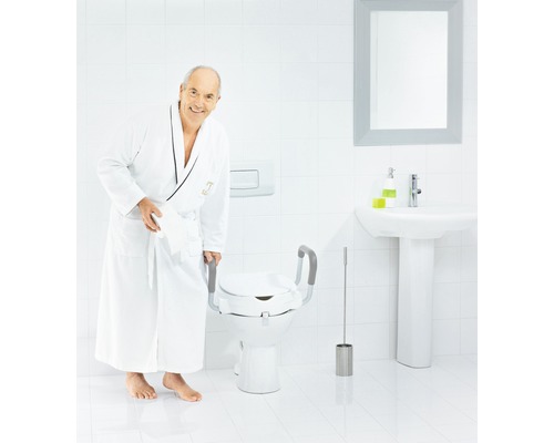 Un bărbat în halat de baie stă lângă o toaletă cu un scaun de toaletă înălțat, inclusiv bare de sprijin.