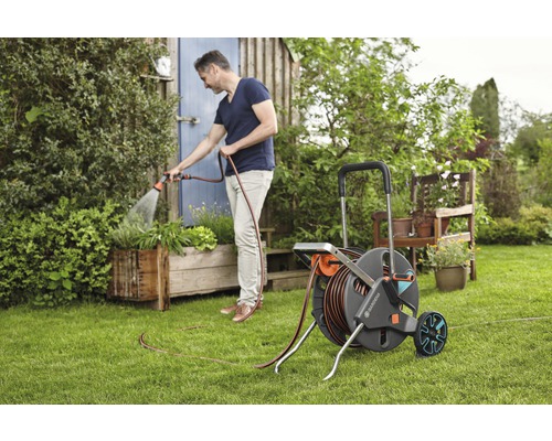 Un bărbat udă plante cu un cărucior cu furtun de grădină în grădină.