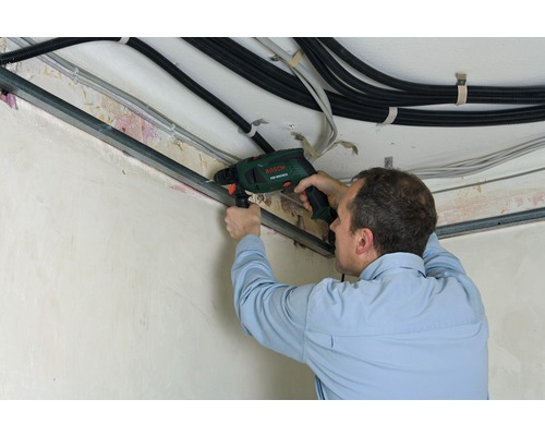 Un bărbat găurește o șină metalică de pe tavan cu o bormașină cu percuție. Cabluri sunt, de asemenea, vizibile.