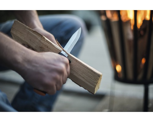 O persoană taie lemne cu un cuțit în fața unui vas pentru foc.