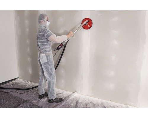 Un bărbat în combinezon de protecție șlefuiește un perete cu o șlefuitoare de tavan.