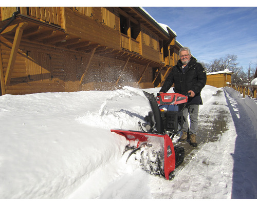 Un bărbat folosește o freză de zăpadă pentru a curăța zăpada în fața unei case de lemn.