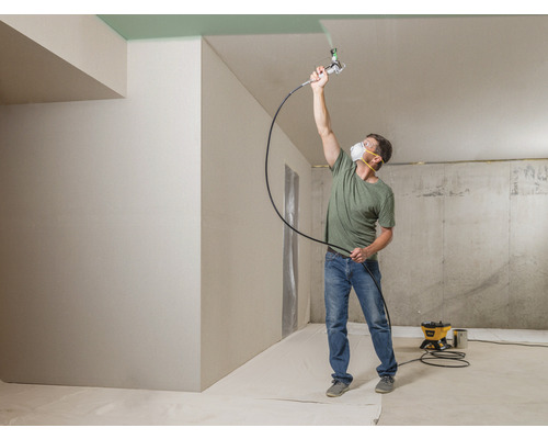 Un bărbat vopsește tavanul cu un pistol de pulverizare a vopselei într-o cameră.