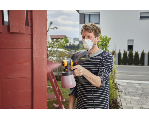 Bărbat vopsește lemn cu sistem de pulverizare a vopselei și mască de protecție