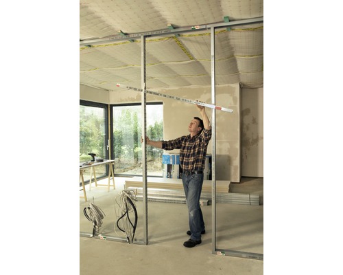 Un bărbat montează un perete despărțitor cu profile metalice într-o construcție nefinisată.