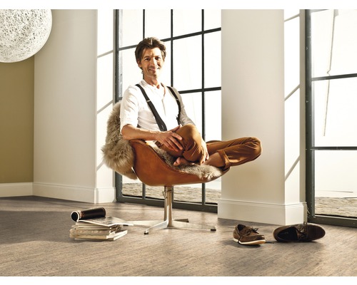 Un bărbat stă relaxat într-un fotoliu într-o cameră luminoasă, cu o fereastră mare și pardoseală.