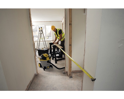 Un muncitor în construcții măsoară cu o ruletă un toc de ușă într-o cameră care necesită renovare. În cameră se află un aspirator, o scară și unelte.