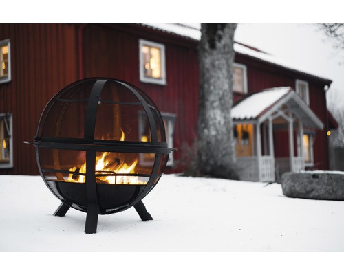 Grătar sferic metalic cu foc în fața unei case roșii în zăpadă