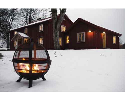 Coș de foc metalic cu foc aprins în grădina înzăpezită din fața unei case.