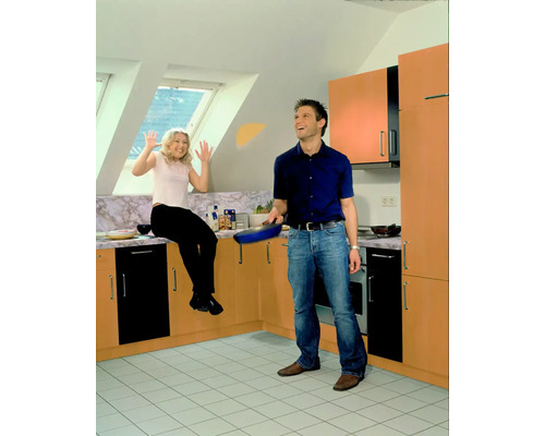 Cameră luminoasă cu un cuplu, bărbat cu tigaie, femeie așezată pe tejgheaua din bucătărie.