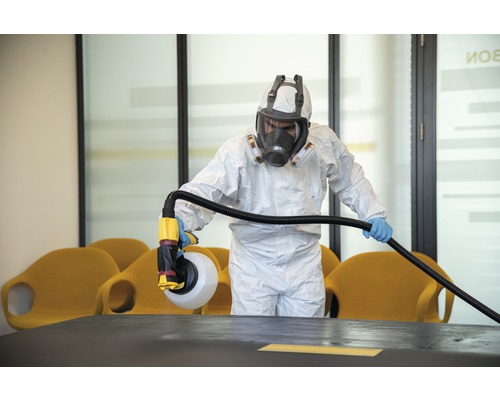 O persoană în echipament de protecție dezinfectează o cameră cu un pulverizator.