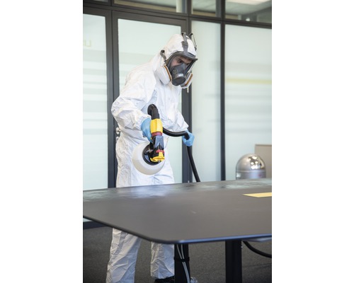 O persoană în echipament de protecție dezinfectează o masă cu un pulverizator de dezinfectant.