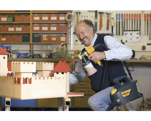 Un bărbat lăcuiește un castel din lemn cu un sistem de pulverizare a vopselei Wagner într-un atelier.