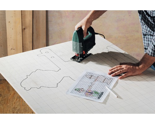 O persoană taie cu un fierăstrău vertical un model dintr-o scândură de lemn. Un șablon și un stilou sunt pregătite.