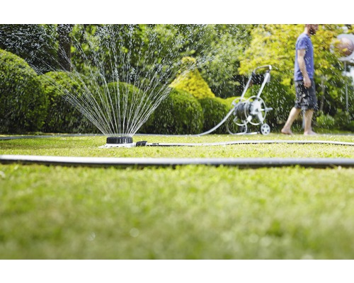 Aspersor de gazon udând un gazon cu un cărucior cu furtun și o persoană în fundal.