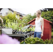 Femeie udând plante cu un furtun de grădină pe o masă de plantare în fața unei căsuțe de grădină.