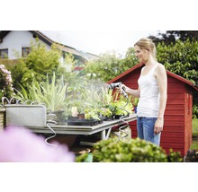 Femeie udând plante cu un pulverizator de grădină pe o masă de grădină în fața unei căsuțe de grădină roșie.