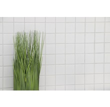 Plantă artificială decorativă în fața unui perete din plăci pătrate albe.