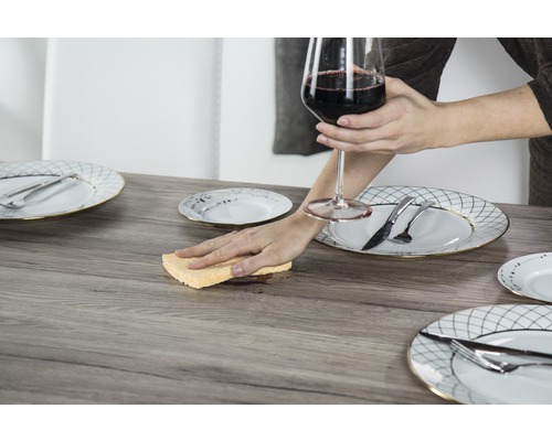 Scenă cu masă, tacâmuri și o persoană care curăță o pată cu un burete în timp ce ține un pahar cu vin.