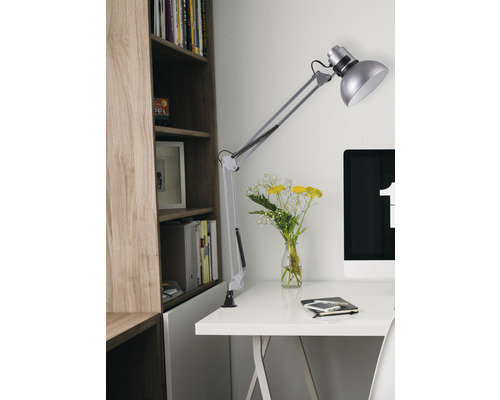 Birou cu lampă cu braț pivotant, computer, caiet și buchet în fața unei biblioteci.