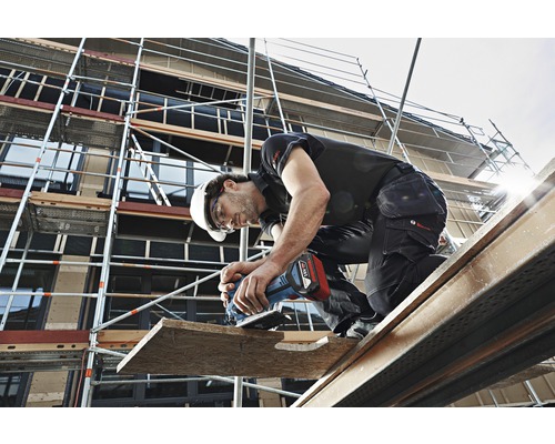 Un muncitor în construcții pe o schelă, care lucrează o scândură de lemn cu o unealtă electrică.