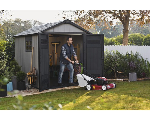 Un bărbat împinge o mașină de tuns iarba dintr-o magazie de grădină într-o grădină.