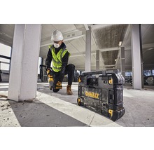 Un muncitor în construcții găurește cu o mașină de găurit într-o clădire, lângă el un încărcător Dewalt.