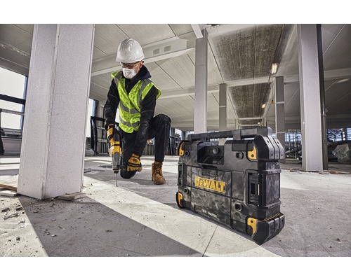 Un muncitor în construcții găurește cu o mașină de găurit într-o clădire, lângă el un încărcător Dewalt.