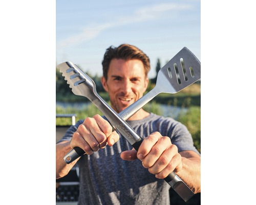 Un bărbat ține un clește de grătar și o spatulă încrucișate în fața camerei.