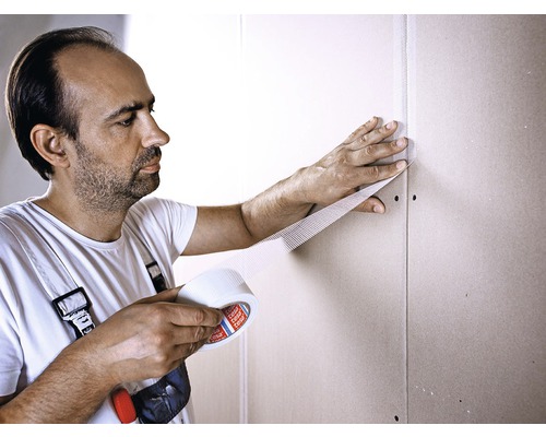 Un bărbat fixează bandă de armare pe un perete din gips-carton.