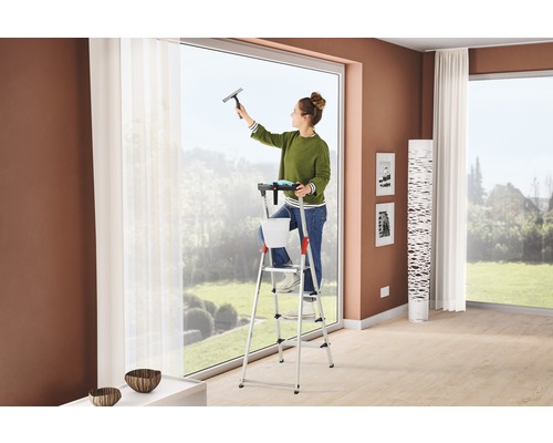 Femeie care curăță ferestrele cu o racletă de geamuri de pe o scară într-un living.