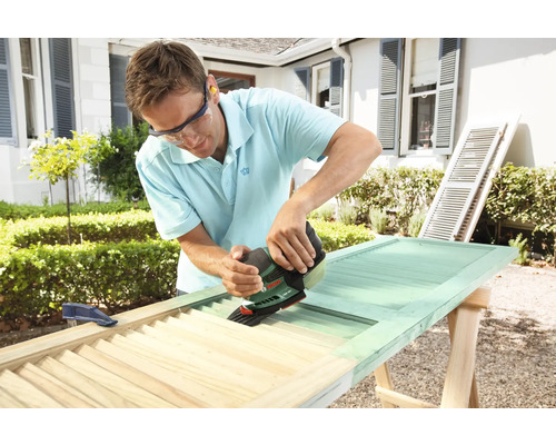 Un bărbat folosește o șlefuitoare pentru a prelucra o jaluzea din lemn. Jaluzeaua se află pe un capră.