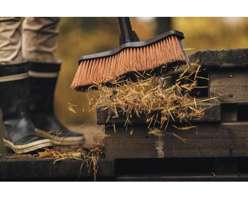 O persoană mătură frunzele și deșeurile de grădină de pe o terasă de lemn cu o mătură de stradă.
