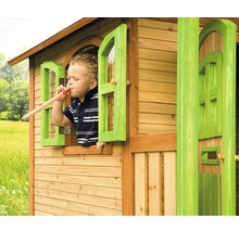 Copil se joacă cu o trompetă într-o căsuță de joacă din lemn cu ferestre verzi.