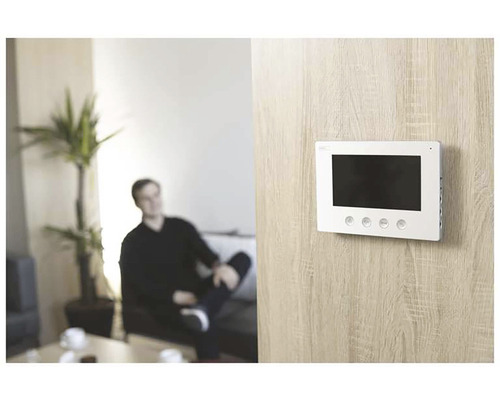 Fotografie de interior a unei sufragerii cu un bărbat pe o canapea și un monitor alb de interfon pe perete.