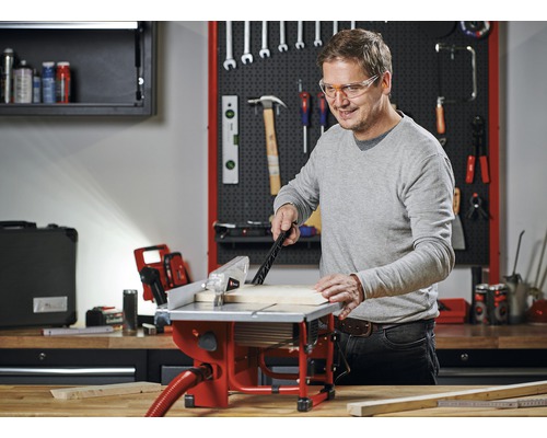 Un bărbat lucrează cu un ferăstrău circular de masă într-un atelier cu scule pe perete.