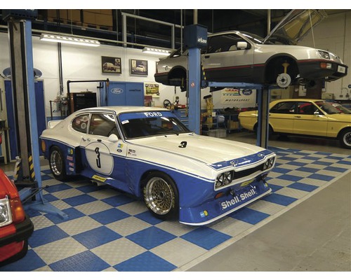 Scenă dintr-un atelier cu mai multe mașini de epocă, inclusiv un Ford Capri cu accente albastre și numărul trei.