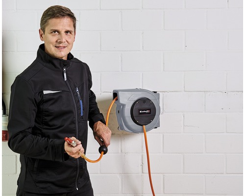 Un bărbat ține un pistol pneumatic pe un tambur de furtun de aer Einhell pe un perete de cărămidă