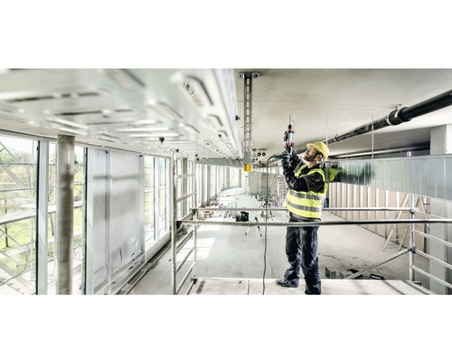 Un muncitor în construcții pe o schelă lucrează cu un ciocan rotopercutor la o instalație de tavan.