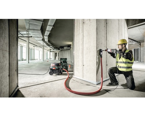 Un muncitor în construcții forează cu un ciocan rotopercutor o gaură într-un perete de beton, aspiratorul de praf este conectat.