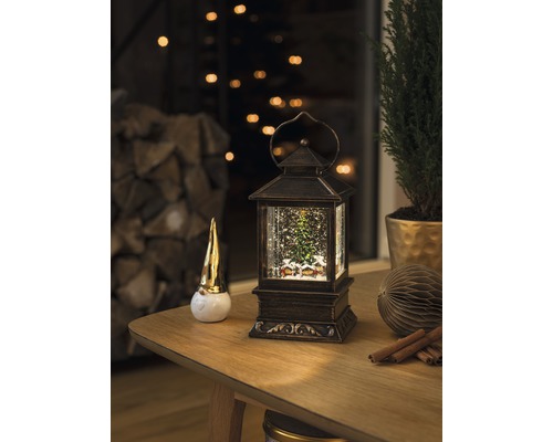 Scenă decorativă cu un felinar glob cu zăpadă, o figurină pitic și o plantă de interior pe o masă