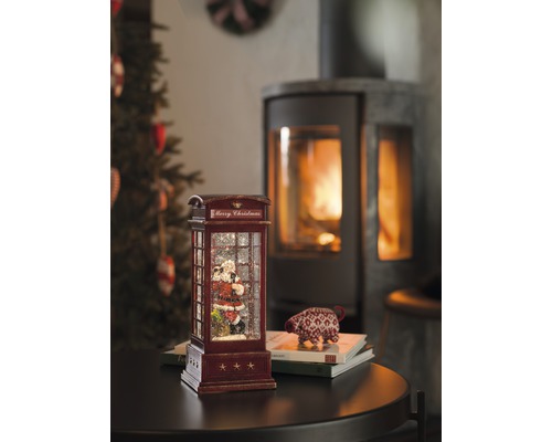 Glob decorativ cu zăpadă în formă de cabină telefonică cu motiv Moș Crăciun și șemineu în fundal.