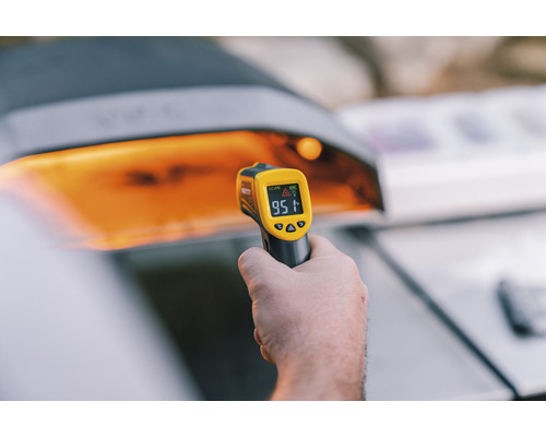 Mână ține un termometru cu infraroșu în fața unui grătar