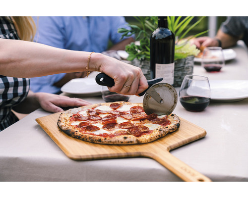 Scenă cu o persoană care taie o pizza cu un tăietor de pizza pe o scândură de lemn, cu o sticlă de vin și pahare în fundal, pe o masă afară