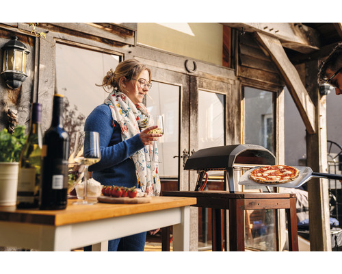 Scenă cu cuptor de pizza, pizza pe o paletă de pizza, masă cu sticle de vin, pahare și o femeie cu un pahar de vin.
