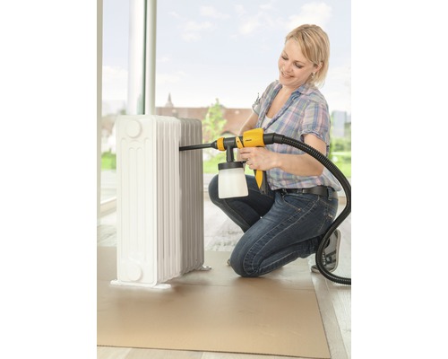 O femeie vopsește un radiator cu un pistol de vopsit într-o cameră luminoasă.