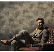 Un bărbat stă așezat în fața unui perete cu tapet cu model circular pe o canapea de piele